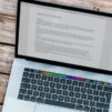 Laptop with text on it with blue/white notebook close to it and pen on notebook. Burgundy flower on the right with phone laying on the wooden table outside