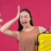 Young Beautiful woman with travel suitcase standing with hand head mistake remember error pink background